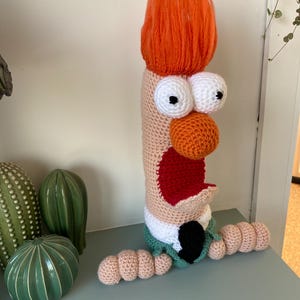 May include: A handmade crochet character with orange hair, large white eyes, and an orange nose. The character has a red mouth, a green collar, and tan arms. The character is sitting on a light blue surface next to green ceramic cacti.