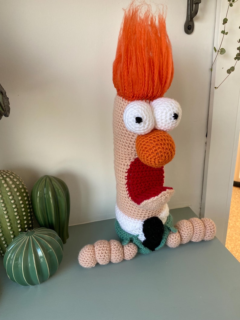 May include: A handmade crochet character with orange hair, large white eyes, and an orange nose. The character has a red mouth, a green collar, and tan arms. The character is sitting on a light blue surface next to green ceramic cacti.
