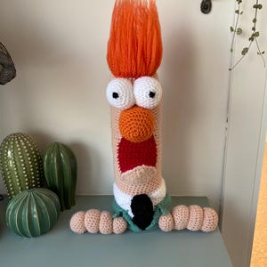 May include: A handmade, crocheted character doll with orange hair, large white eyes, and an orange nose. The doll has a red mouth, a green jacket, and a black tie. The doll's arms are made of crocheted, rounded shapes. The doll is standing on a blue-green surface.