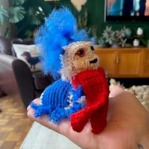 May include: A small, handcrafted crochet creature featuring a fuzzy, bright blue tuft of hair, a cream-colored face with dark eyes, and a blue body. The figure is holding a vibrant red, textured, rectangular piece of fabric in its front limbs.
