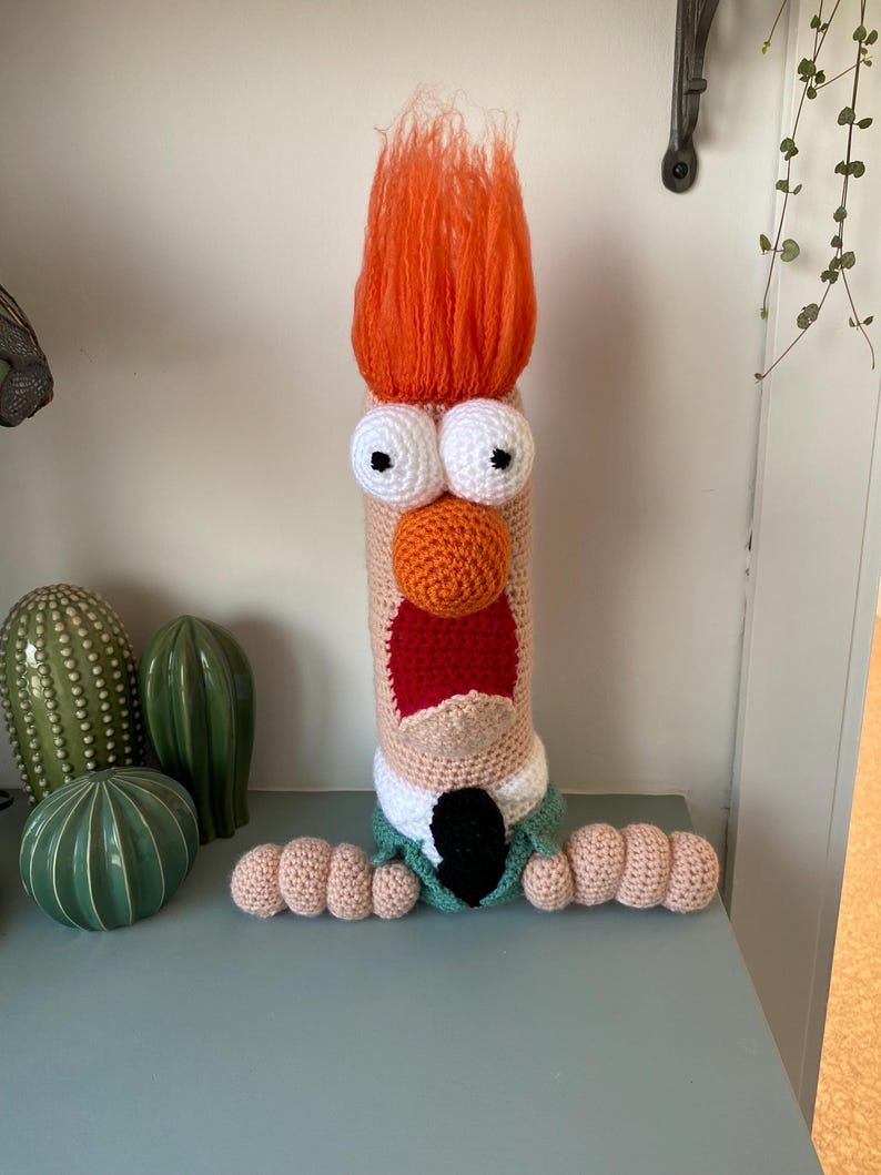 May include: A handmade, crocheted character doll with orange hair, large white eyes, and an orange nose. The doll has a red mouth, a green jacket, and a black tie. The doll's arms are made of crocheted, rounded shapes. The doll is standing on a blue-green surface.