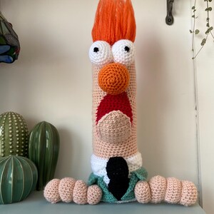 May include: A handmade, crocheted character with orange hair, white eyes, and an orange nose. The character has a light skin tone, a red mouth, and is wearing a green jacket with a black tie. The character is a whimsical, decorative item.