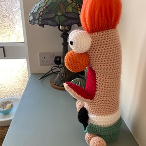 May include: A whimsical, handmade crochet creature with a peach-colored body, orange hair, and a white and orange eye. It has a red and black detail and a segmented tail. The creature is displayed on a light blue surface.
