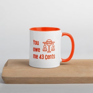 May include: White ceramic mug with an orange rim and handle. The mug has a red graphic of a scale with a dollar bill on top, a female symbol on one side, and a male symbol on the other side. The text "You owe me 43 cents" is printed below the graphic.