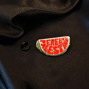 Puede incluir: Un pin de esmalte en forma de sandía con las palabras "FREE PALESTINE" en letras rojas. El pin tiene un borde dorado, una corteza verde y semillas negras. Un pasador negro está al lado del pin.