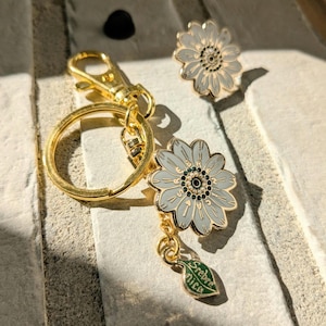May include: A gold-colored keychain with a daisy charm and a matching daisy pin. The daisy charms are light gray with a gold outline and a black and green center. A green leaf charm with the text "Srebrnica" hangs from the keychain.