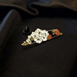 Könnte beinhalten: Ein floraler Emaille-Ansteckpin mit einem Bouquet aus weißen Blumen, goldenen Akzenten und roten Blüten auf schwarzem Hintergrund. Der Pin hat einen goldenen Rand und einen schwarzen Nadelverschluss. Das Design ist detailliert und elegant.