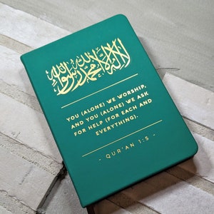 Könnte beinhalten: Ein rechteckiges, smaragdgrünes Buch mit goldener arabischer Kalligraphie und englischem Text. Der Text lautet: "YOU (ALONE) WE WORSHIP, AND YOU (ALONE) WE ASK FOR HELP (FOR EACH AND EVERYTHING)." Das Buch trägt den Titel "QUR'AN 1:5".