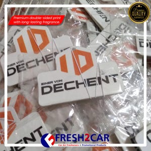 May include: White car air fresheners with an orange and white logo that reads "ID DECHENT". The text "Premium double-sided print with long-lasting fragrance" is also visible. The fresheners are packaged in clear plastic.