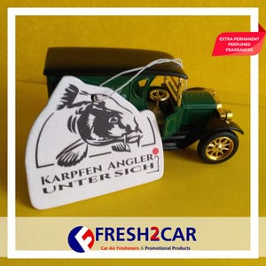 May include: A car air freshener with the text "KARPFEN ANGLER UNTER SICH" and a carp illustration, hanging in front of a green and black vintage car model. The freshener is white with black text and a black illustration. The car model has gold wheels.