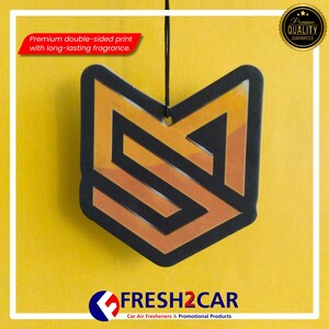 Pu&ograve; includere: Un deodorante per auto con un design geometrico nero e arancione, appeso su uno sfondo giallo. Il testo "Premium double-sided print with long-lasting fragrance" &egrave; visibile. Il logo "FRESH2CAR" &egrave; in basso.