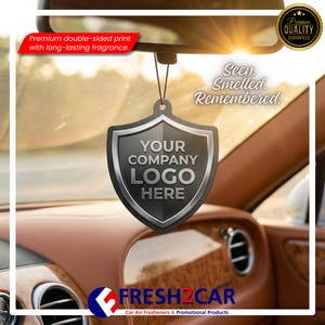 May include: A black shield-shaped car air freshener with the text "YOUR COMPANY LOGO HERE" hangs from a car's rearview mirror. The text "Seen. Smelled. Remembered." is visible. The product is from FRESH2CAR, a car air freshener and promotional products company.