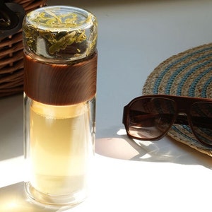 Op de afbeelding: Een glazen theefilterfles met een houten deksel en een bruine houten hoes. De fles is gevuld met losse thee en de thee is zichtbaar door het glas.