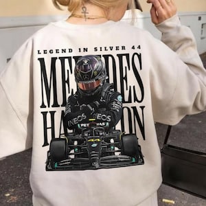 Peut inclure: Sweat-shirt crème avec un graphisme de voiture de course de Formule 1 et un pilote. Le texte "LEGEND IN SILVER 44" et "MERCEDES HAMILTON" sont imprimés au dos. Le pilote porte un casque avec le logo Petronas.