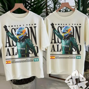 Puede incluir: Dos camisetas color crema con temática de Fórmula 1. Cada camiseta presenta un gráfico de un piloto con traje y casco de carreras verdes, con las palabras "ASTON ALONSO" en letras negras grandes. También es visible el número "14".