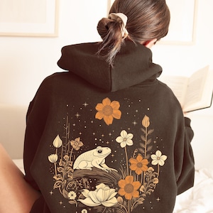 May include: A dark brown hoodie featuring a graphic print of a frog sitting in a field of flowers. The flowers are in shades of orange, yellow, and white. The background is a dark blue with white stars.