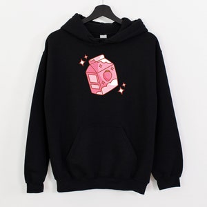 May include: Black hoodie with a pink cartoon illustration of a milk carton with a strawberry on top. The carton has a white lid and a white cloud on the side. There are small white stars around the carton.