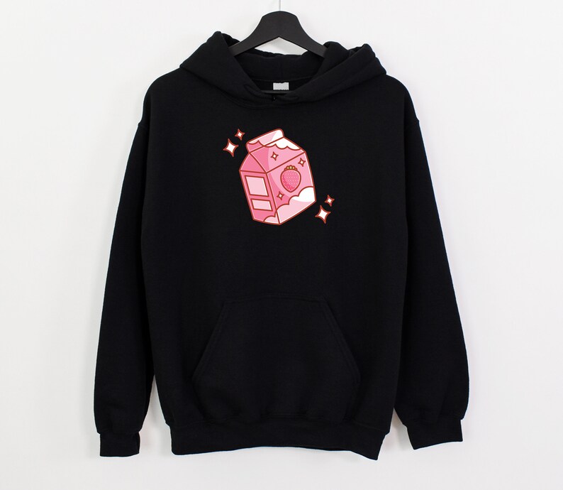 May include: Black hoodie with a pink cartoon illustration of a milk carton with a strawberry on top. The carton has a white lid and a white cloud on the side. There are small white stars around the carton.