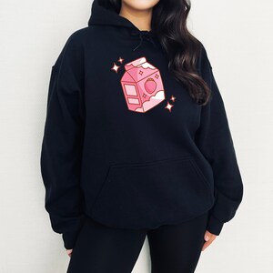 May include: A black hoodie with a pink cartoon illustration of a milk carton with a strawberry on it. The carton has a white lid and a white cloud with stars above it.