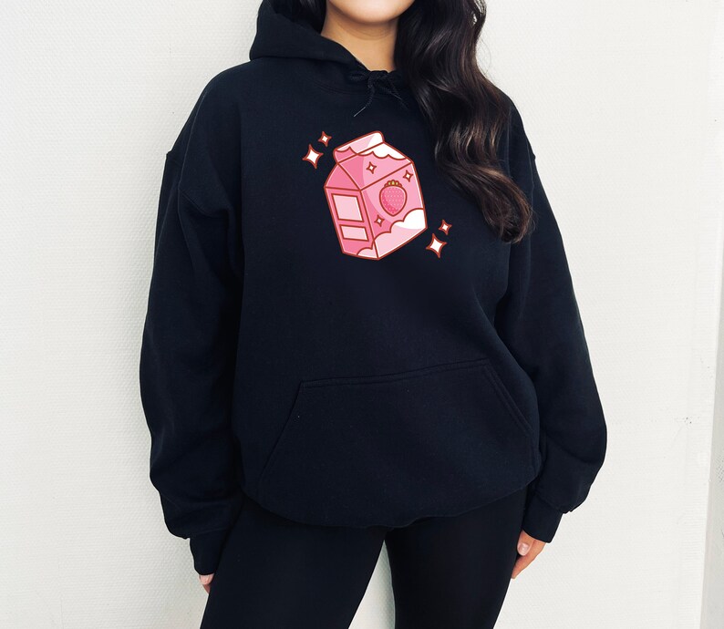 May include: A black hoodie with a pink cartoon illustration of a milk carton with a strawberry on it. The carton has a white lid and a white cloud with stars above it.
