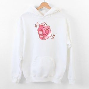 May include: White hoodie with a pink cartoon illustration of a milk carton with a strawberry on top. The carton has a pink lid and a white cloud with a pink outline. There are two pink stars on either side of the carton.