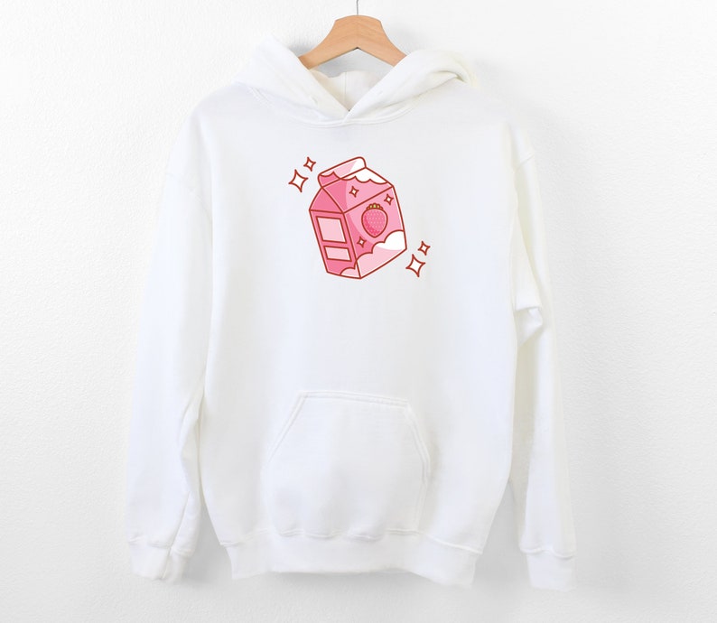 May include: White hoodie with a pink cartoon illustration of a milk carton with a strawberry on top. The carton has a pink lid and a white cloud with a pink outline. There are two pink stars on either side of the carton.