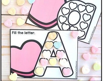 Op de afbeelding: Een printbare Valentijnsdagactiviteit voor kinderen. De afbeelding toont een werkblad met de letter A die zwart is omlijnd. De letter is gevuld met roze, gele en blauwe snoephartjes. De tekst "Vul de letter" staat bovenaan het werkblad. Het werkblad ligt op een houten oppervlak met verspreide snoephartjes.
