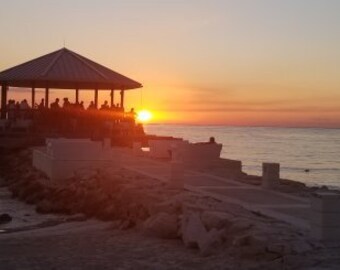 May include: A group of people are gathered under a wooden gazebo on a pier overlooking the sea at sunset. The sky is a vibrant orange and pink, and the water is calm and blue.
