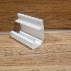 May include: A clear, plastic clip with a curved base and a slotted top section. The clip is designed to hold items and is shown on a wooden surface.