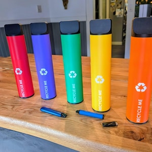 Può includere: Cinque oggetti cilindrici colorati con coperchi neri e la scritta "RECYCLE ME" e un simbolo di riciclaggio. Gli oggetti sono rossi, viola, verdi, gialli e arancioni. Due oggetti blu e un oggetto nero sono alla base.