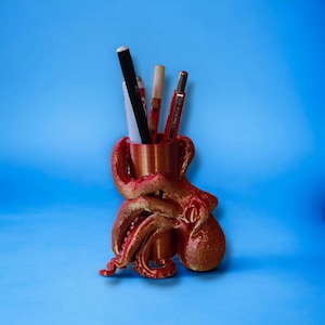 May include: A red and gold octopus-shaped desk organizer holding pens and pencils. The tentacles of the octopus wrap around a cylindrical cup. The organizer is made of a textured material and sits on a blue background.