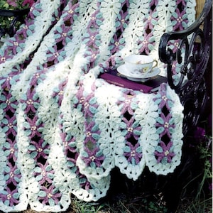 May include: A crocheted blanket with a floral pattern in shades of white, purple, and green. The blanket is draped over a black metal bench with a book and a teacup on top.