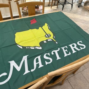 May include: Green flag featuring the Masters Tournament logo. The logo includes a yellow outline of a golf course with a red flag and the word "MASTERS" in white. The flag is displayed on a wooden table.