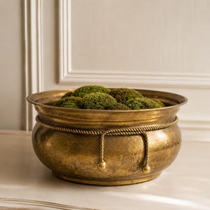 May include: A round, antique brass planter filled with vibrant green moss. The planter has a wide rim and is encircled by a decorative rope detail with hanging tassels. The aged brass finish gives it a vintage look, suitable for home decor.