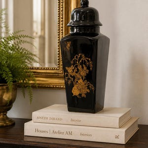 May include: A glossy black ceramic ginger jar with a matching lid, featuring a square base and tapered sides. The jar is decorated with intricate gold-colored illustrations of a bird perched among flowering branches and butterflies.