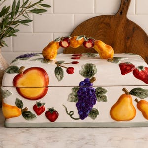 May include: A rectangular ceramic bread box with a white base featuring raised, colorful fruit motifs including apples, strawberries, grapes, and pears. The lid has a sculpted handle shaped like a cluster of fruit, including small apples and pears.