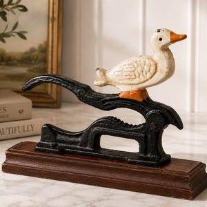May include: A cast iron nutcracker featuring a white duck with an orange beak mounted on a black metal lever mechanism. The entire assembly is attached to a rectangular dark brown wooden base, creating a functional tool for cracking nuts.
