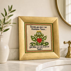 May include: A framed cross-stitch artwork featuring a green frog with a red heart, wearing a crown. The text reads, "Before you meet the handsome prince, you have to kiss a lot of toads!" The frame is gold.
