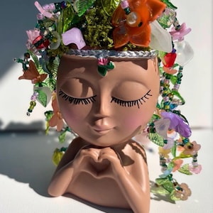 May include: A terracotta-colored head-shaped planter with closed eyes and long eyelashes, featuring hands forming a heart shape. The head is adorned with a crown of colorful glass beads, green leaves, and orange flowers. The text Gabrielle is below.