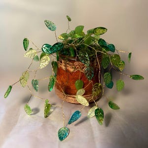 May include: A decorative plant sculpture featuring numerous translucent green and teal glass leaves attached to thin, flexible gold-toned wire branches. The wire stems are anchored in a small, cylindrical brown pot wrapped in textured twine.