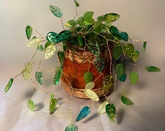 May include: A decorative plant sculpture featuring numerous translucent green and teal glass leaves attached to thin, flexible gold-toned wire branches. The wire stems are anchored in a small, cylindrical brown pot wrapped in textured twine.
