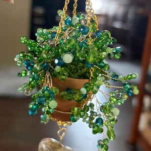 May include: Miniature hanging planter with a terracotta pot filled with green and blue beaded foliage. The planter is suspended by a gold chain and features a clear, heart-shaped charm. A charming, handcrafted decorative item.