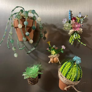 Może przedstawiać: Kolekcja magnetycznych roślin i kwiatów z koralików. Magnesy obejmują wiszącą roślinę w doniczce z terakoty, bukiet z kokardą, małą kompozycję kwiatową, sukulent w plecionym koszu i kaktus w doniczce.