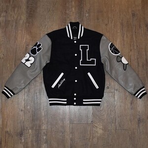 Puede incluir: Chaqueta universitaria negra con mangas grises. Presenta una gran "L" blanca en el pecho, un parche de huella de pata y "22" en una manga, y un parche de baloncesto y personaje de dibujos animados en la otra. El nombre "Ashley" está bordado en la parte delantera.