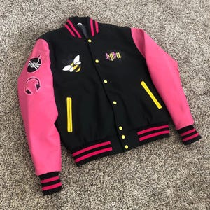 May include: Black varsity jacket with pink sleeves, featuring a bee and music-themed patches. The jacket has yellow accents, the word "AyeBee" in script, and red and black striped cuffs and hem.
