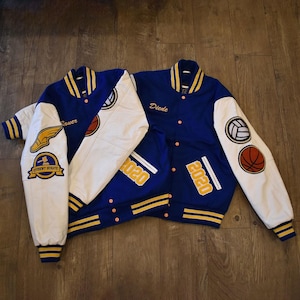 Puede incluir: Dos chaquetas universitarias azules y blancas con detalles dorados. Una chaqueta tiene el nombre "Rauer" y un parche de "Student Senate". La otra chaqueta tiene el nombre "Diede" y el año "2020" en la espalda. Ambas chaquetas tienen parches deportivos.