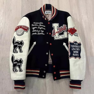 Può includere: Una giacca college blu navy e bianca con dettagli rossi. La giacca presenta toppe con testo e illustrazioni di gatti sulle maniche. Il testo include "Lone Wolf", "Ruf-Tuf" e "For Toughest Leather Work Boots". Il colletto e i polsini hanno dettagli a righe.