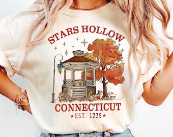 May include: A cream colored t-shirt with a graphic of a gazebo with fall foliage and the text "Stars Hollow Connecticut *EST. 1779*"