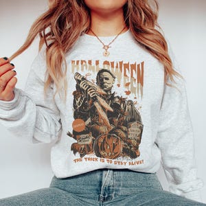 May include: A light gray Halloween-themed sweatshirt featuring a graphic of Michael Myers holding a knife, pumpkins, and the text "HALLOWEEN". The phrase "THE TRICK IS TO STAY ALIVE!" is printed at the bottom.