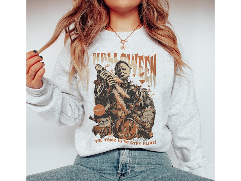 May include: A light gray Halloween-themed sweatshirt featuring a graphic of Michael Myers holding a knife, pumpkins, and the text "HALLOWEEN". The phrase "THE TRICK IS TO STAY ALIVE!" is printed at the bottom.